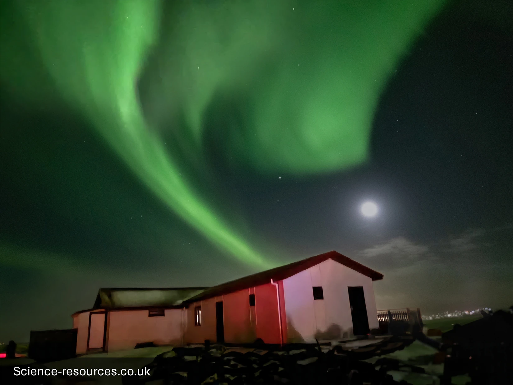 A night-time photograph taken in Iceland in March 2025, showing vivid green aurora borealis sweeping across the sky in wide, flowing arcs. The aurora forms a twisting, luminous ribbon that curves above a small, single‑storey house with a red roof and pale exterior walls. The house sits in a dark, rocky landscape illuminated faintly by the glow of the aurora and a bright full moon on the right. Stars are visible through gaps in the auroral light, and thin clouds hover near the horizon. The scene feels remote and peaceful, dominated by the striking natural light display overhead.