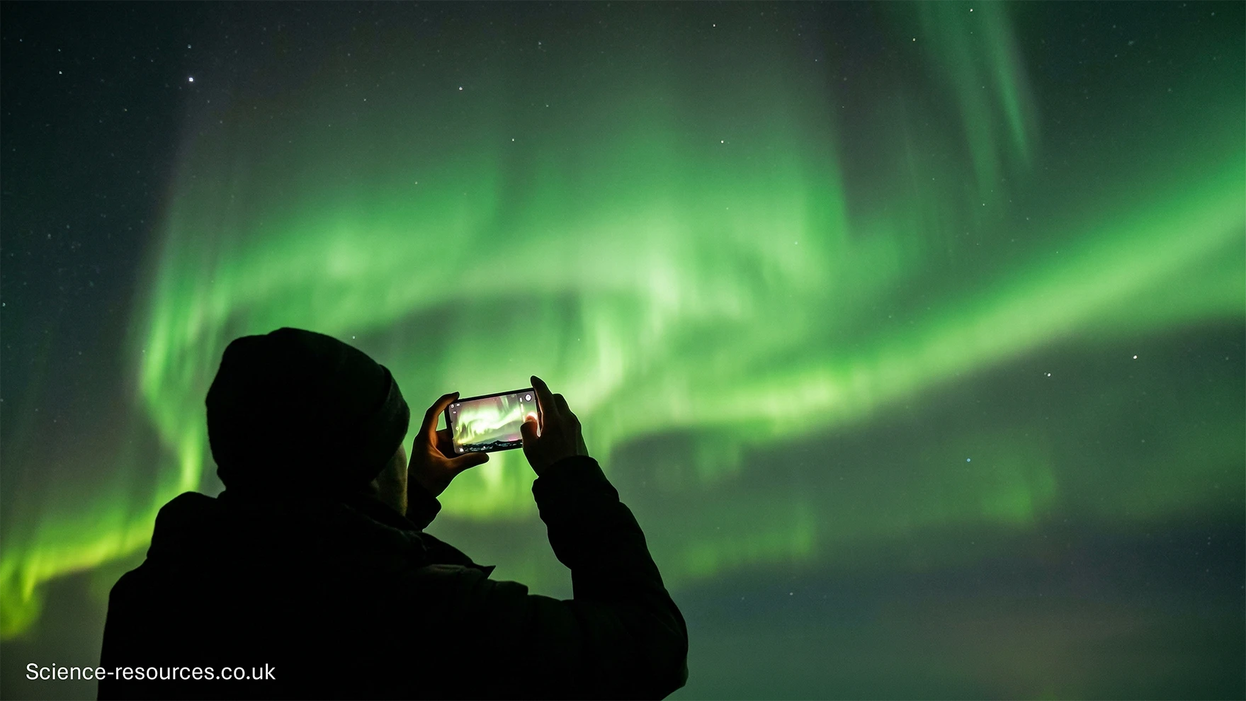The image shows a silhouetted person holding up a mobile phone to capture a bright, vivid display of the Northern Lights. The aurora stretches across the sky in sweeping bands of green light, with subtle variations in brightness and form. The person appears to be outdoors at night, bundled against the cold, focusing their phone camera on the glowing sky above. The overall scene conveys a sense of wonder and the accessibility of photographing aurora displays using everyday devices.